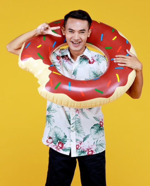 Hombre asiático atractivo joven con sombrero y camisa hawaiana blanca con estampados de flores rojas y verdes y anillo de natación donut alrededor de su cuello para vacaciones en la playa sobre fondo amarillo.