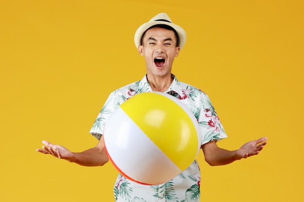 Hombre asiático atractivo joven con sombrero y camisa hawaiana blanca con estampados de flores rojas y verdes sosteniendo una pelota de playa amarilla con ir de vacaciones en la playa sobre fondo amarillo.