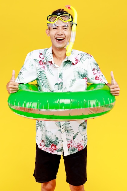 Hombre asiático atractivo joven con sombrero y camisa hawaiana blanca con máscara amarilla de snorkel y anillo de natación verde alrededor de su cintura sobre fondo amarillo. Concepto de vacaciones en la playa.
