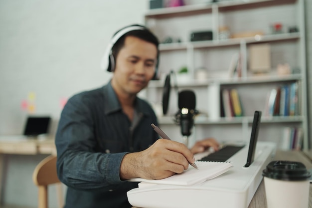 Un hombre asiático con auriculares blancos toca el piano y canta una canción y aprende en línea con un teléfono móvil y compone una canción o graba sonido con un micrófono y usa un programa de grabación de música por computadora