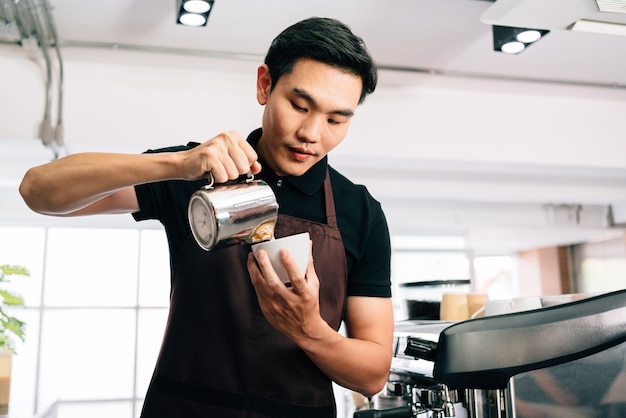 Hombre asiático barista en un delantal, vertió intencionalmente leche caliente en un café negro espresso caliente.