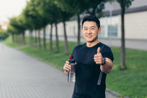 Hombre asiático bebe agua después del entrenamiento físico y trotar hombre feliz sonríe muestra el pulgar hacia arriba y mira a la cámara