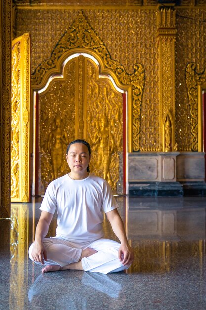 El hombre asiático de cabello largo relaja la meditación con todos los disfraces blancos sentados frente al papel tapiz dorado de Buddist en el Templo, Tailandia.