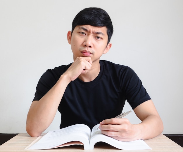 Hombre asiático camisa negra cara seria y tocar su barbilla con el libro sobre el escritorio mirar a la cámara