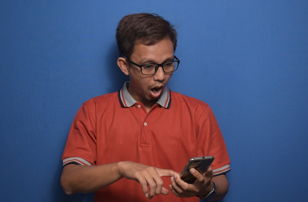 Hombre asiático con camiseta casual naranja con teléfono inteligente con una cara de sorpresa y expresión de asombro
