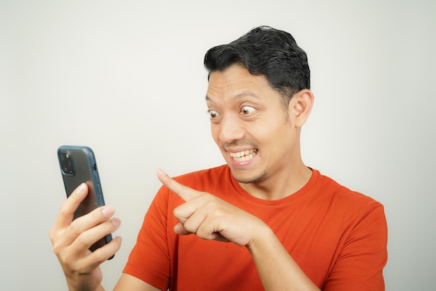 Hombre asiático con camiseta naranja enojado mirando el mensaje en su teléfono inteligente en un fondo aislado