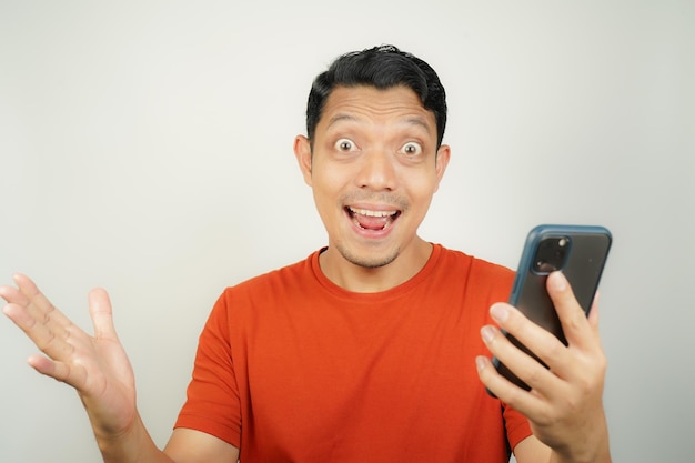 Hombre asiático con camiseta naranja feliz mirando lo que en el teléfono inteligente en un fondo aislado