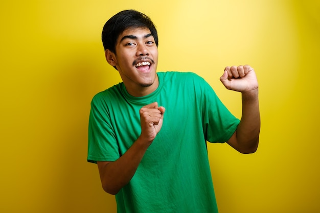 Hombre asiático en camiseta verde sonriendo y bailando alegremente, expresando alegremente celebrando buenas noticias victoria ganando gesto de éxito contra el fondo amarillo