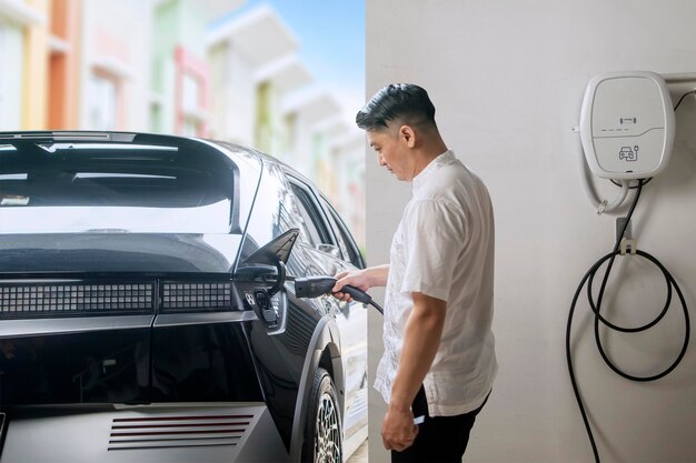 Hombre asiático cargando su moderno vehículo eléctrico en el garaje de su casa