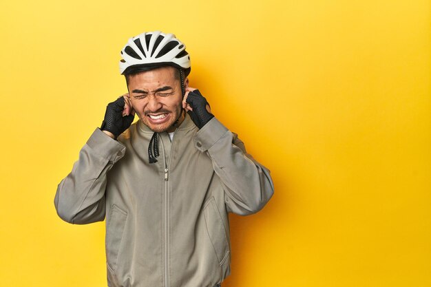 Hombre asiático con casco de bicicleta y guantes telón de fondo de estudio amarillo cubriendo las orejas con las manos