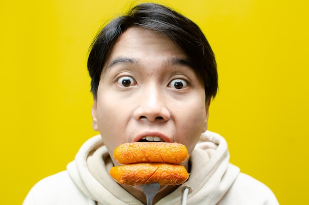 hombre asiático come y sostiene una salchicha en un tenedor aislado sobre un fondo amarillo de comida rápida poco saludable