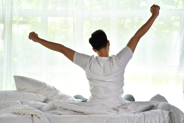 Hombre asiático despertando por la mañana sentado en la cama y estirando