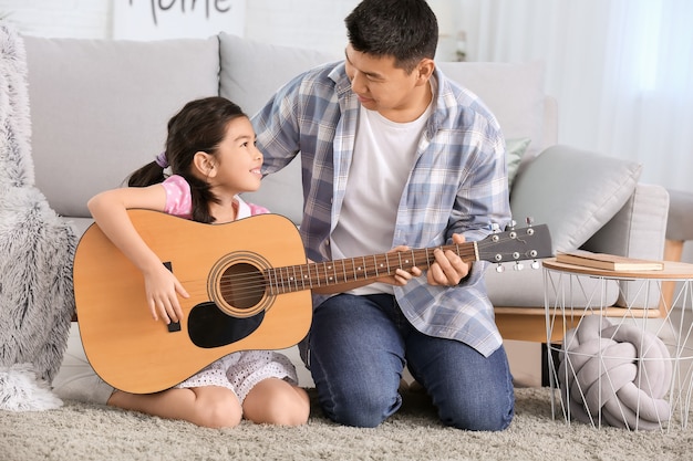 Hombre asiático enseñando a su pequeña hija a tocar la guitarra en casa