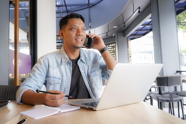 Un hombre asiático está haciendo una llamada telefónica con camisa azul usando una computadora portátil y un teléfono móvil en una cafetería