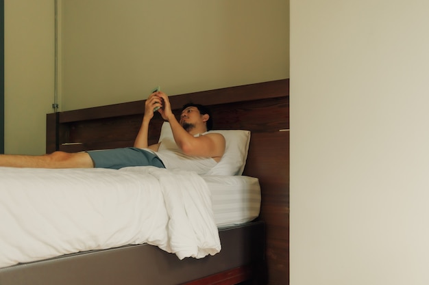 Hombre asiático está usando smartphone en la cama en el hotel. Concepto de pereza y relax.