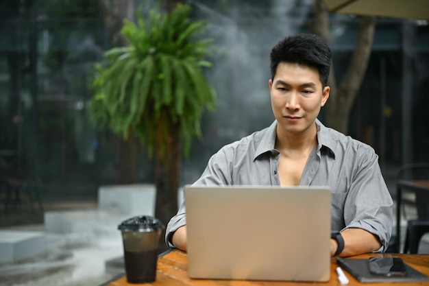 Un hombre asiático está usando su computadora portátil trabajando en su trabajo y sentado en una cafetería.