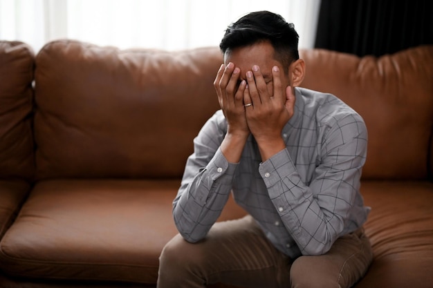 Un hombre asiático estresado y deprimido se cubre la cara llorando en el sofá de una sala de estar