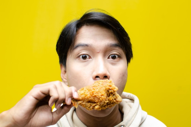 Un hombre asiático extático y expresivo come y muerde pollo frito aislado sobre un fondo amarillo.