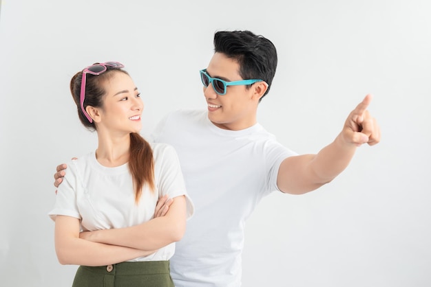 Hombre asiático feliz abrazando a novia sonriente mientras señala con el dedo aislado en blanco