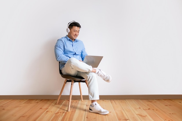 Hombre asiático feliz en auriculares con videollamada en la computadora portátil sentado en una silla contra la pared blanca espacio libre de cuerpo entero