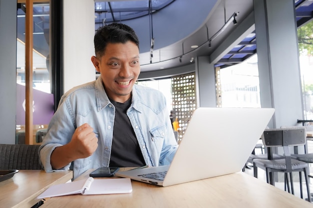 Hombre asiático feliz y entusiasta con camisa azul usando portátil y teléfono móvil en el negocio de compras en línea de la cafetería