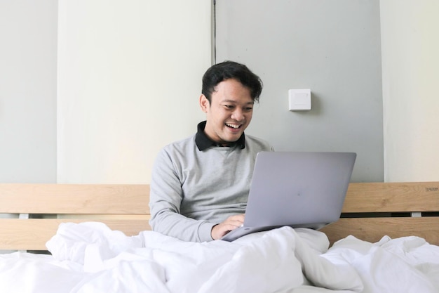 Hombre asiático feliz está trabajando con su computadora portátil en su cama Concepto de estilo de vida exitoso independiente