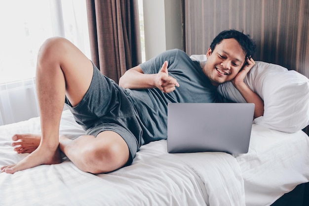 Hombre asiático feliz está trabajando con su computadora portátil en su cama Concepto de estilo de vida exitoso independiente