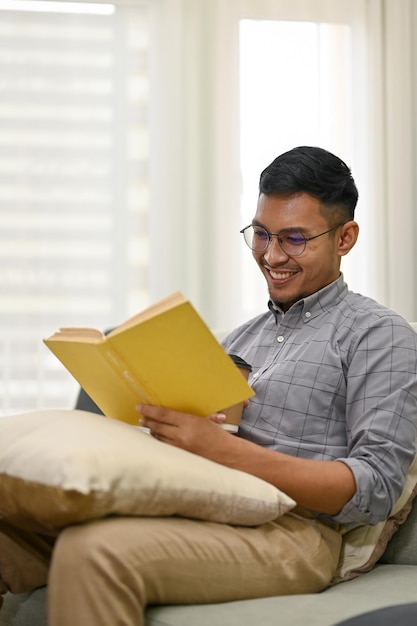 Hombre asiático feliz leyendo un libro en el sofá en la sala de estar