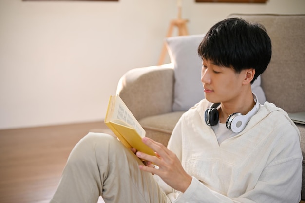 Hombre asiático feliz leyendo un libro en su sala de estar mínima disfruta leyendo novela