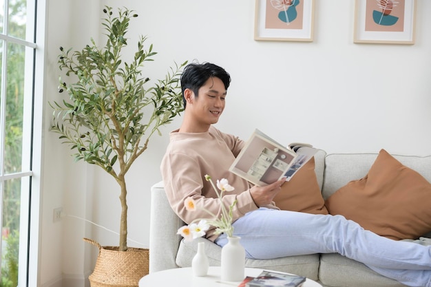 Hombre asiático feliz sentado en el sofá leyendo un libro en la sala de estar en casa