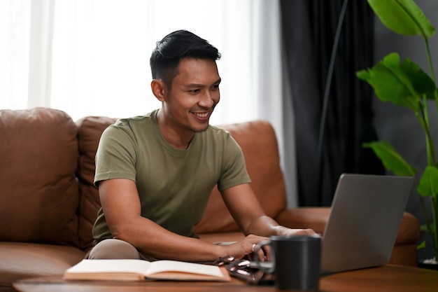 Hombre asiático feliz usando una computadora portátil en un sofá en la sala de estar trabajando buscando en línea