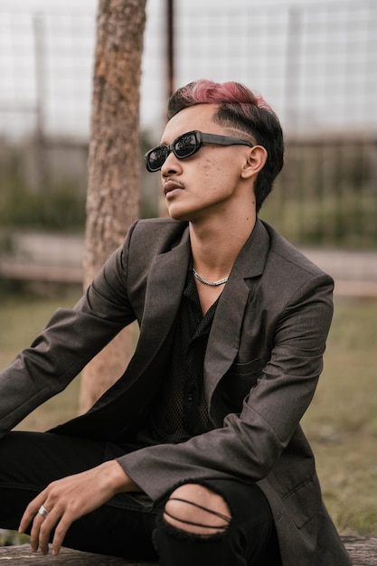 Un hombre asiático con gafas de sol está sentado en el tronco de un árbol con un estilo muy masculino en el patio