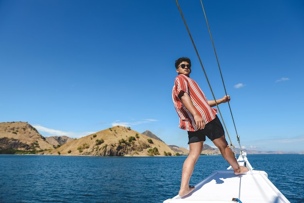 Hombre asiático en gafas de sol posando en el borde del barco con paisaje marino y colinas en Labuan Bajo