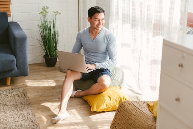 Hombre asiático gay guapo que trabaja con una computadora portátil sentada cerca de la ventana en la sala de estar en casa Hombre LGBT guapo sonriendo usando una computadora portátil y mirando fuera de la ventana Concepto de vida LGBT