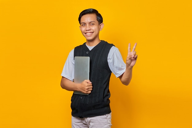 Hombre asiático guapo sosteniendo portátil sonriendo y mostrando el signo de la paz sobre fondo amarillo