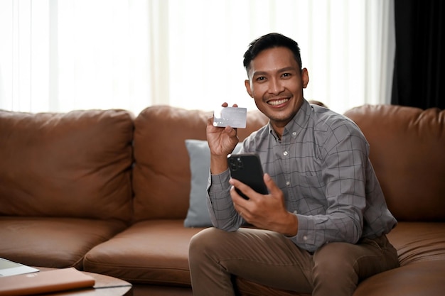 Hombre asiático guapo sosteniendo su teléfono inteligente y mostrando una tarjeta de crédito a la cámara