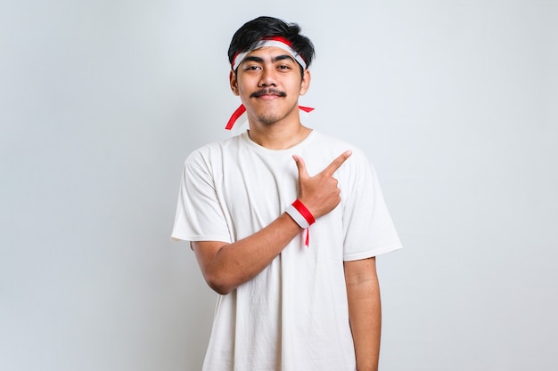 Hombre asiático hermoso joven que lleva la venda roja y blanca sobre el fondo blanco con una gran sonrisa en la cara; apuntando con el dedo de la mano hacia el lado mirando a la cámara.