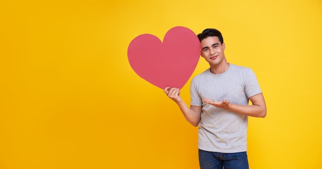 Hombre asiático hermoso joven que muestra la muestra del corazón rojo en amarillo. amor y feliz concepto de San Valentín.