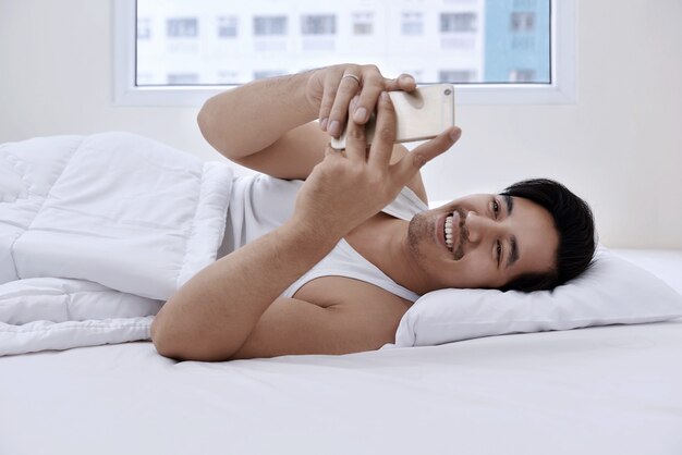 Hombre asiático hermoso que miente en cama con smartphone