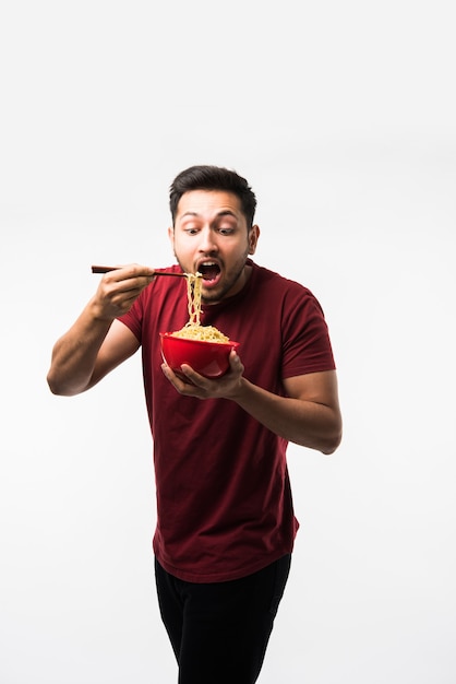 Hombre asiático indio comiendo fideos calientes o ramen en un tazón rojo con palillos