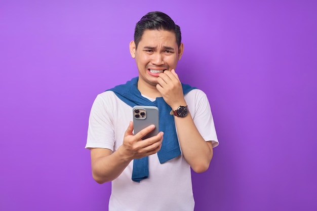 Hombre asiático joven asustado en camiseta casual sosteniendo teléfono móvil reaccionando a malas noticias aislado sobre fondo púrpura Concepto de estilo de vida de la gente