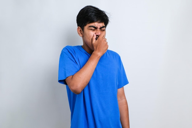 Hombre asiático joven con camisa casual sobre fondo blanco que huele algo apestoso y desagradable, un olor intolerable, conteniendo la respiración con los dedos en la nariz. Mal olor