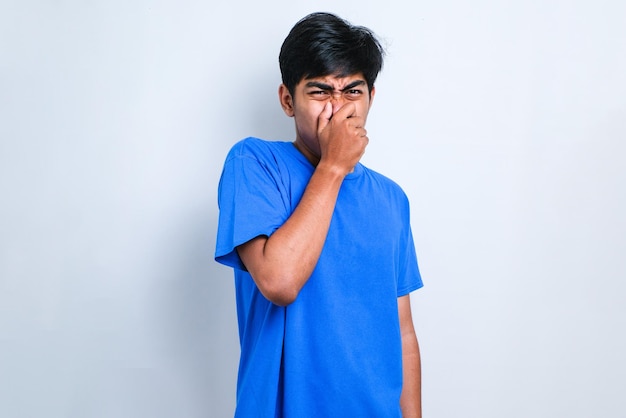 Hombre asiático joven con camisa casual sobre fondo blanco que huele algo apestoso y desagradable, un olor intolerable, conteniendo la respiración con los dedos en la nariz. Mal olor