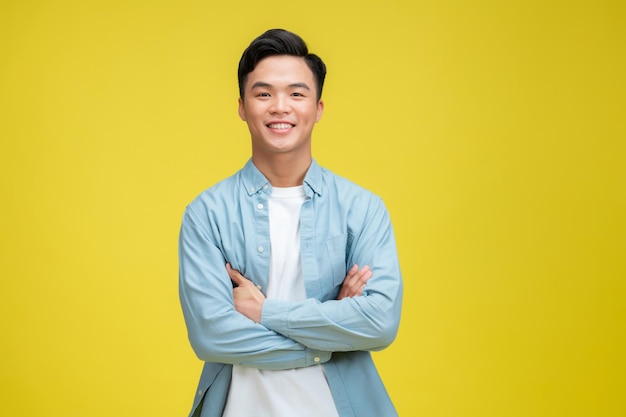 Hombre asiático joven confiado que se coloca de brazos cruzados contra fondo amarillo