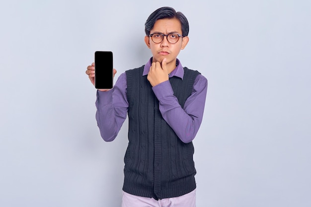 Hombre asiático joven confundido en camisa casual y chaleco que muestra la pantalla en blanco del teléfono móvil recomendando la aplicación aislada sobre fondo blanco Concepto de estilo de vida de la gente