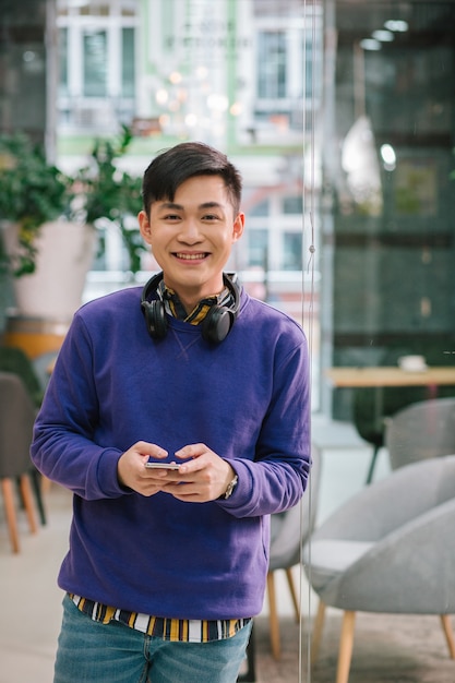 Hombre asiático joven contento de pie solo con un teléfono inteligente moderno en sus manos y sonriendo. Habitación con puerta de vidrio a sus espaldas