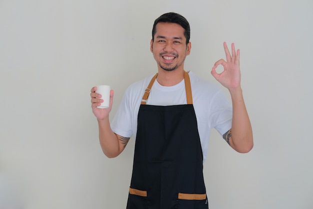 Hombre asiático joven con delantal de barista sonriendo amable mientras sostiene la taza de bebida