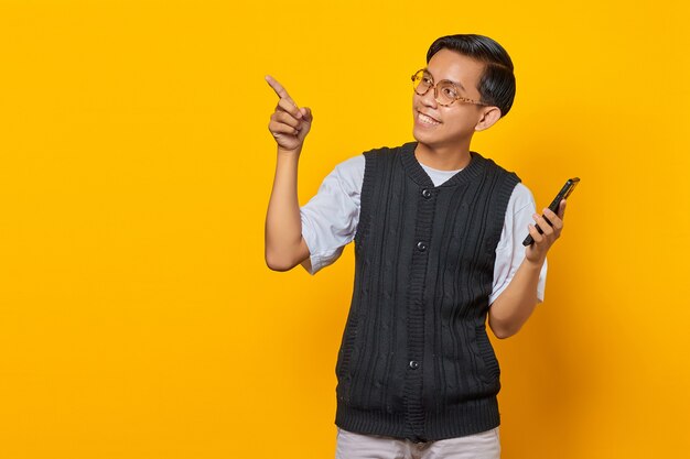 Hombre asiático joven feliz que sostiene el teléfono móvil y que señala el dedo para copiar el espacio en fondo amarillo