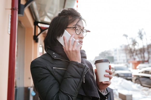 Hombre asiático joven hermoso que habla en el teléfono mientras bebe café.