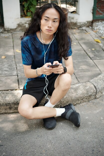 Hombre asiático joven hermoso serio con el pelo rizado sentado en la acera y escuchando un mensaje de audio en auriculares al aire libre